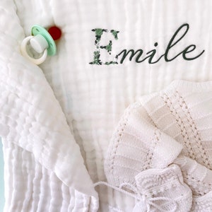 Pu&ograve; includere: Coperta per neonato in mussola bianca con una lettera E ricamata con fiori verdi e il nome "Emile" in ricamo nero. Sono visibili anche un berretto per neonato in maglia bianca e un ciuccio.