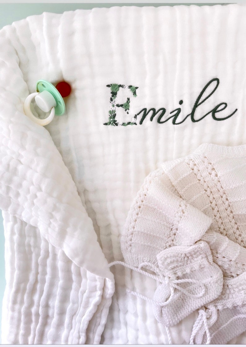 Pu&ograve; includere: Coperta per neonato in mussola bianca con una lettera E ricamata con fiori verdi e il nome "Emile" in ricamo nero. Sono visibili anche un berretto per neonato in maglia bianca e un ciuccio.