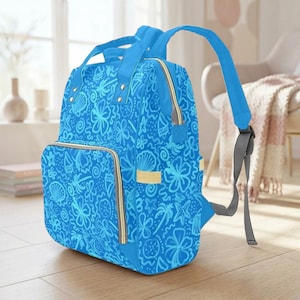 Peut inclure: Un sac à dos bleu avec un motif tropical de fleurs, coquillages et palmiers. Il possède une poche avant, une poignée supérieure et des bretelles réglables. Les fermetures éclair sont dorées.