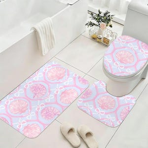 May include: A three-piece bathroom set featuring a seashell pattern. The set includes a rectangular bath mat, a U-shaped toilet mat, and a toilet seat cover. The design features pink seashells and white bows on a light blue background. The set is designed to coordinate.
