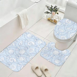 May include: A light blue bathroom set featuring a seashell and rope pattern. The set includes a rectangular bath mat, a U-shaped toilet mat, and a toilet seat cover. White slippers are in the foreground.