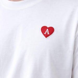 Puede incluir: Camiseta blanca con un parche rojo en forma de corazón. El parche en forma de corazón tiene un borde blanco y una letra "A" blanca en el centro. La camiseta es de un material suave, probablemente algodón.