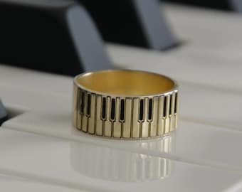 Puede incluir: Un anillo dorado con un diseño de teclas de piano descansa sobre las teclas blancas de un piano. El anillo presenta una banda de teclas de piano en miniatura, alternando negro y dorado. Las teclas del piano son negras y blancas.