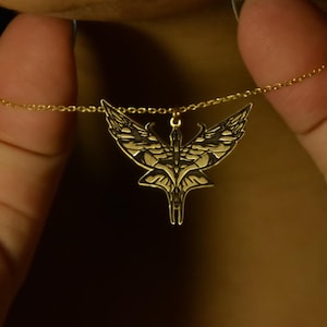 May include: A gold-colored necklace with a pendant shaped like stylized wings. The pendant is suspended from a delicate chain. The necklace is held between two hands against a dark background.