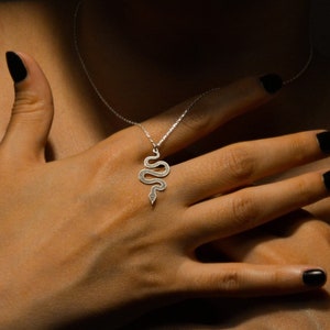 May include: A silver snake pendant necklace. The pendant is a stylized snake design with a delicate chain. The person in the image is wearing a black top and has black nail polish.