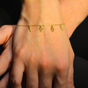 May include: A gold bracelet with four small charms is displayed on a wrist. The charms include a key, a circle, a bottle, and a lightning bolt. The person's nails are painted a light purple colour.