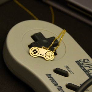 Pode incluir: Um colar com pingente dourado com um design de controle de videogame. O colar é exibido em um controle Super Nintendo vintage, com cartuchos de jogos ao fundo. O controle é cinza com botões pretos.
