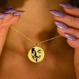 May include: A gold-colored pendant necklace featuring a circular charm with a black stylized mask design. The necklace is held by a person with long, polished nails. The chain is delicate and thin, and the pendant is centered.