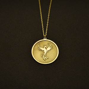 May include: Gold-colored pendant necklace featuring a phoenix design within a circular frame. The pendant hangs from a delicate chain. The necklace is set against a dark background, highlighting the jewelry's details.