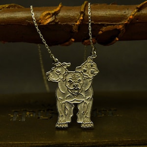 May include: A silver-colored pendant necklace featuring a three-headed dog design. The dog has a fierce expression and is suspended from a delicate chain. The necklace is displayed against a dark background.