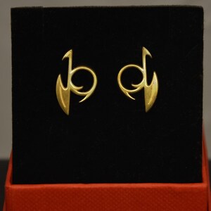 Puede incluir: Un par de pendientes de oro presentados sobre una superficie de terciopelo negro dentro de una caja roja. Cada pendiente presenta un diseño estilizado con un elemento curvo y circular y una forma puntiaguda en forma de ala.