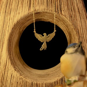 May include: A gold-colored pendant necklace featuring a phoenix design. The necklace is displayed against a textured, wooden background with a circular cutout. Two decorative bird figurines are in the foreground.