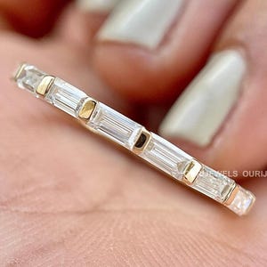 May include: A gold band ring with a row of baguette-cut diamonds set in a continuous line around the band. The ring is on a finger.
