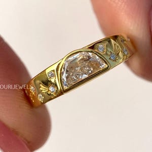 May include: A gold ring with a half-moon shaped clear stone and small diamond accents. The ring has a floral design and the brand name "OURI JEWELS" is visible.