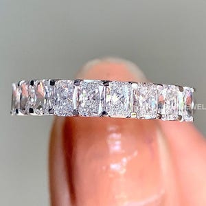 May include: A silver band ring with a row of square-shaped diamonds. The ring is on a finger.