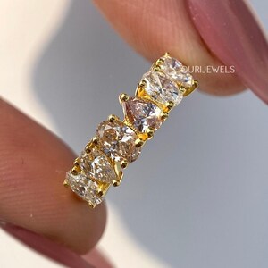 May include: A gold ring featuring a series of clear and blush-colored pear-shaped gemstones. The ring is held between fingers with a pink nail polish. The brand name "OURI JEWELS" is visible.