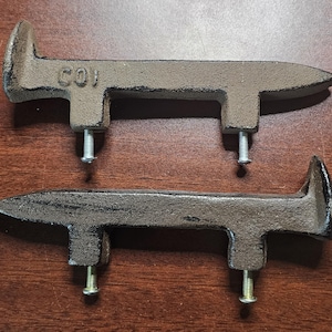 Op de afbeelding: Twee gietijzeren kastgrepen met een rustieke, verweerde uitstraling. Elke greep heeft een T-vormig ontwerp met een puntig uiteinde en een montageschroef. Het woord "COI" is zichtbaar op een van de grepen.