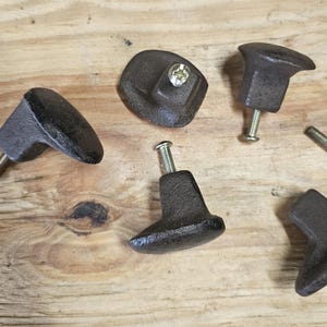 May include: Five dark brown cast iron cabinet knobs with screws. The knobs have a rustic, textured appearance and various geometric shapes. The knobs are arranged on a light brown wooden surface.