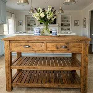 May include: A rustic wooden kitchen island featuring two drawers with metal pulls and two slatted lower shelves. A clear glass vase filled with white flowers and two blue and white patterned ceramic canisters sit on the top surface.