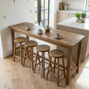 May include: A rectangular wooden bar table with four matching round wooden stools. The table surface holds three ceramic cups on saucers and a ceramic teapot. The furniture features a natural wood grain finish and is set on a light wood floor.