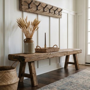 May include: A rustic wooden bench with a thick, weathered plank seat and four sturdy legs. On top sits a ceramic jug filled with dried wheat stalks, a woven rectangular tray with handles, and a tall, thin, dark brown taper candle in a holder.