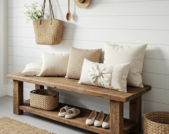 May include: A wooden entryway bench with a lower shelf, featuring a wall-mounted coat rack with five black hooks. The bench holds four cream-colored pillows, including one with a bow detail. A woven tote bag, a hat, and a wooden spoon hang on the hooks.