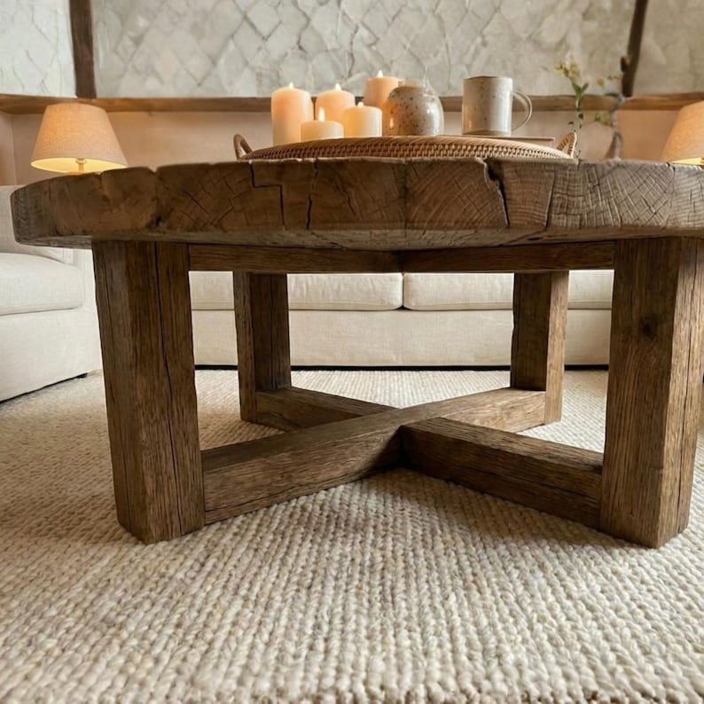 May include: A round coffee table constructed from thick, reclaimed brown wood with a visible grain and a cross-base support structure. On the tabletop sits a woven tray containing several pillar candles and a ceramic mug.