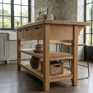 May include: A light brown wooden kitchen island featuring three top drawers, a central open shelf holding stacked bowls, and a bottom shelf containing a dark ceramic jar and a woven basket. Two stools with tan leather seats are tucked underneath.