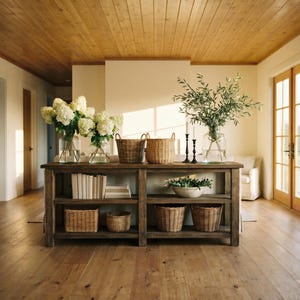 May include: A rustic wooden console table displays home decor items. It features glass vases with white flowers and greenery, woven baskets, books, and candles. The room has a wooden ceiling and floor, with natural light.