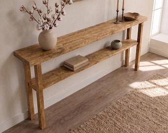 May include: A long, narrow wooden console table with a natural light brown finish, featuring a rectangular top and a lower shelf. The table is supported by four straight legs and holds a vase with cotton stems, two candles, and a small bowl.
