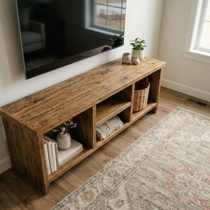 May include: A long, rectangular wooden media console with a natural brown wood grain finish. The unit features three open storage compartments. The left section holds white books and a small vase, the center holds a folded textile, and the right holds a woven basket.