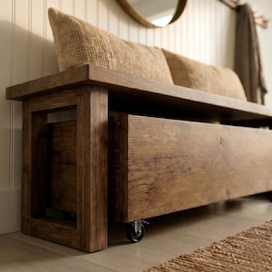 May include: A rectangular wooden bench with a dark brown finish and visible wood grain. The bench features a lower storage compartment equipped with small black metal caster wheels for mobility. Two textured beige cushions rest on the seat.