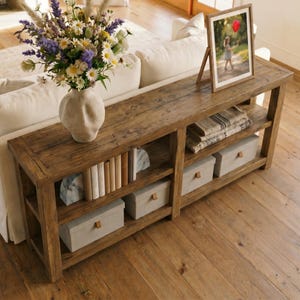 May include: A long, rectangular wooden console table with a rustic brown finish, featuring two open shelves. The top surface holds a textured off-white vase with a wildflower bouquet and a framed photograph. Lower shelves contain books and storage boxes.