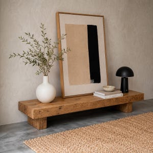 May include: A rustic wooden bench with a framed abstract art piece, a white vase with greenery, a black table lamp, and a small bowl on a stack of books. A woven jute rug is in the foreground.