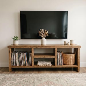 May include: A rustic, wooden TV stand with open shelving, displaying vinyl records, a woven basket, and decorative items. A large black television is mounted above the stand. A small plant and vase with dried flowers add to the decor.