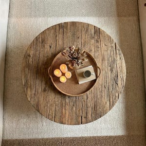May include: A round, weathered wooden coffee table featuring a textured surface with visible grain. Centered on the table is a round woven tray containing four glowing pillar candles, a small ceramic mug, and a dried floral arrangement.