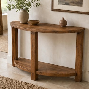 May include: A semi-circular wooden console table with a natural brown finish, featuring a flat top surface and a lower shelf supported by four vertical legs. A beige ceramic vase with green foliage and a small brown bowl sit on the top surface.