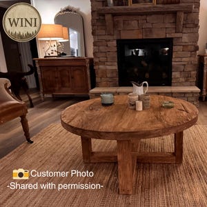 May include: A round, medium-brown wooden coffee table with a thick, circular top and a sturdy cross-base support structure. The table features a visible wood grain texture and is positioned on a woven rug, displaying a natural, unfinished appearance.