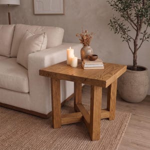 May include: A square wooden side table with a natural grain finish and four sturdy legs connected by a cross-brace base. The tabletop holds two cream-colored pillar candles, a ceramic vase with dried flowers, a small wooden bowl, and books.