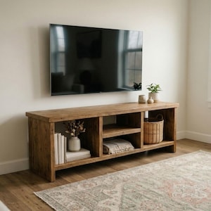 May include: A rustic, wooden TV stand with open shelving and a woven basket. The stand is made of light brown wood and has a natural, unfinished look. A large, flat-screen TV is mounted above the stand. The room has a neutral color palette.
