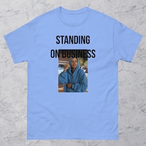 May include: Light blue t-shirt with the text "STANDING ON BUSINESS" in black. The shirt features a small image of a person wearing a blue hoodie. The person is making a hand gesture.