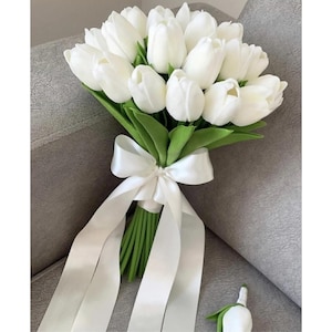 以下が含まれることがあります： 白いチューリップのブーケ。緑の葉と白いサテンのリボンで結ばれています。ブーケにはお揃いのブートニアも含まれています。花は灰色の表面に配置されています。