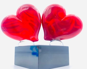 May include: Two red glass hearts with swirling patterns, sitting on a black square base.