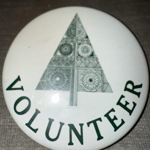 May include: White button with a green geometric tree design and the word "VOLUNTEER" in green lettering. The tree is composed of various geometric shapes and patterns. The button is round and has a glossy finish.