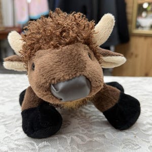 Puede incluir: Un peluche de bisonte con pelaje marrón suave, un mechón de pelo rizado en la cabeza, cuernos de color crema y pezuñas negras. El animal de peluche tiene una nariz lisa de color gris oscuro y ojos pequeños de color negro, en posición erguida.