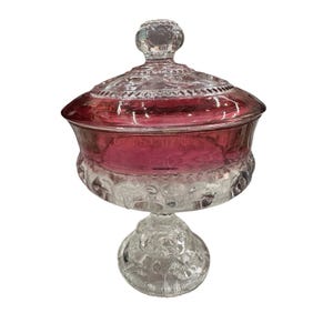 Puede incluir: Un bombonera de cristal transparente con interior y tapa de color rojo rubí. El plato tiene una base de pedestal y un diseño decorativo texturizado. La tapa presenta un pomo redondo y un patrón detallado. Es un artículo decorativo.