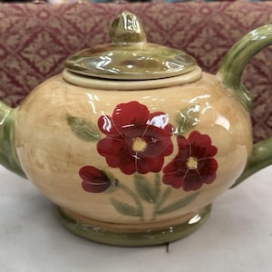 May include: A ceramic teapot with a round body, a lid, and a curved handle. The teapot is painted with a beige background and red floral designs. The spout and handle are green.