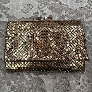 May include: A vintage gold mesh clutch bag with a rectangular shape. The bag features a clasp closure and is made of small, circular metal pieces. The background is a white lace fabric.