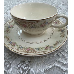 Può includere: Servizio da tè color crema con decorazioni floreali e bordi dorati. La tazza ha un manico curvo e poggia su un piattino abbinato. Il set presenta un delicato motivo floreale rosa, blu e verde.