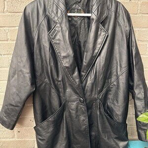 May include: A black leather jacket with a single button closure and two large pockets. The jacket has a collar and a long, flowing style.